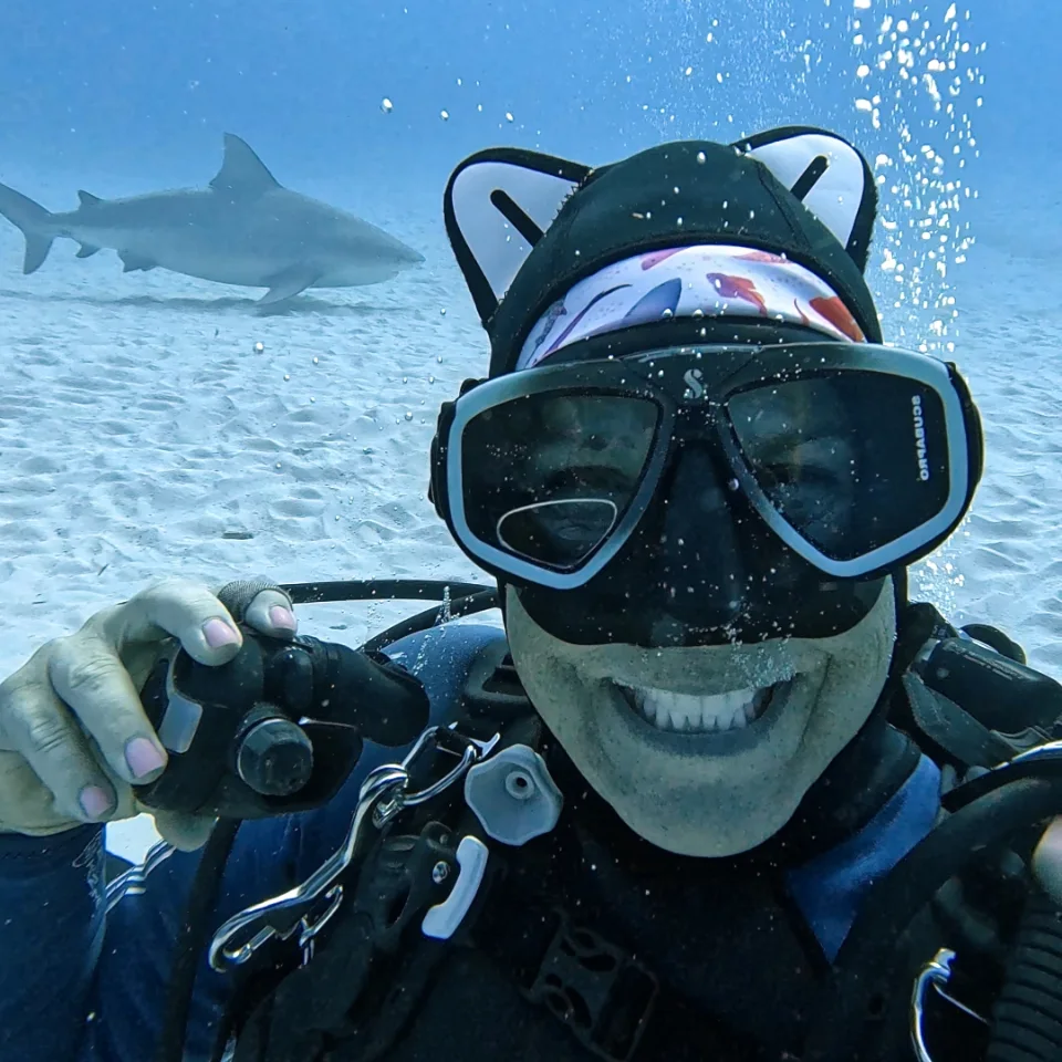 Diver smiling at the camera with a bull shark passing behind