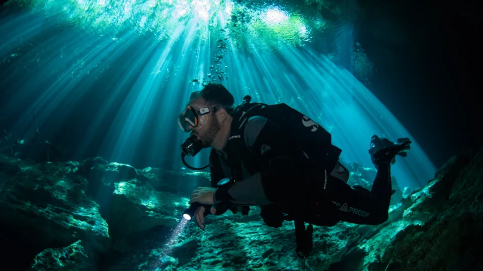 Diver with a torch gliding beneath cenote light beams near Puerto Aventuras